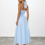 A woman modeling a light blue halter-neck maxi dress from the back, featuring a ruched bodice, tie-back detail, and a flowing skirt, set against a plain white wall and gray floor.