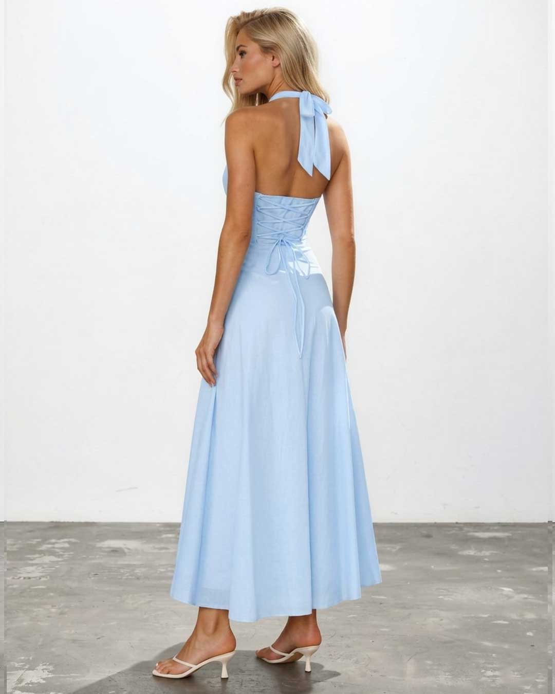 A woman modeling a light blue halter-neck maxi dress from the back, featuring a ruched bodice, tie-back detail, and a flowing skirt, set against a plain white wall and gray floor.
