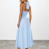 A woman modeling a light blue halter-neck maxi dress from the back, featuring a ruched bodice, tie-back detail, and a flowing skirt, set against a plain white wall and gray floor.