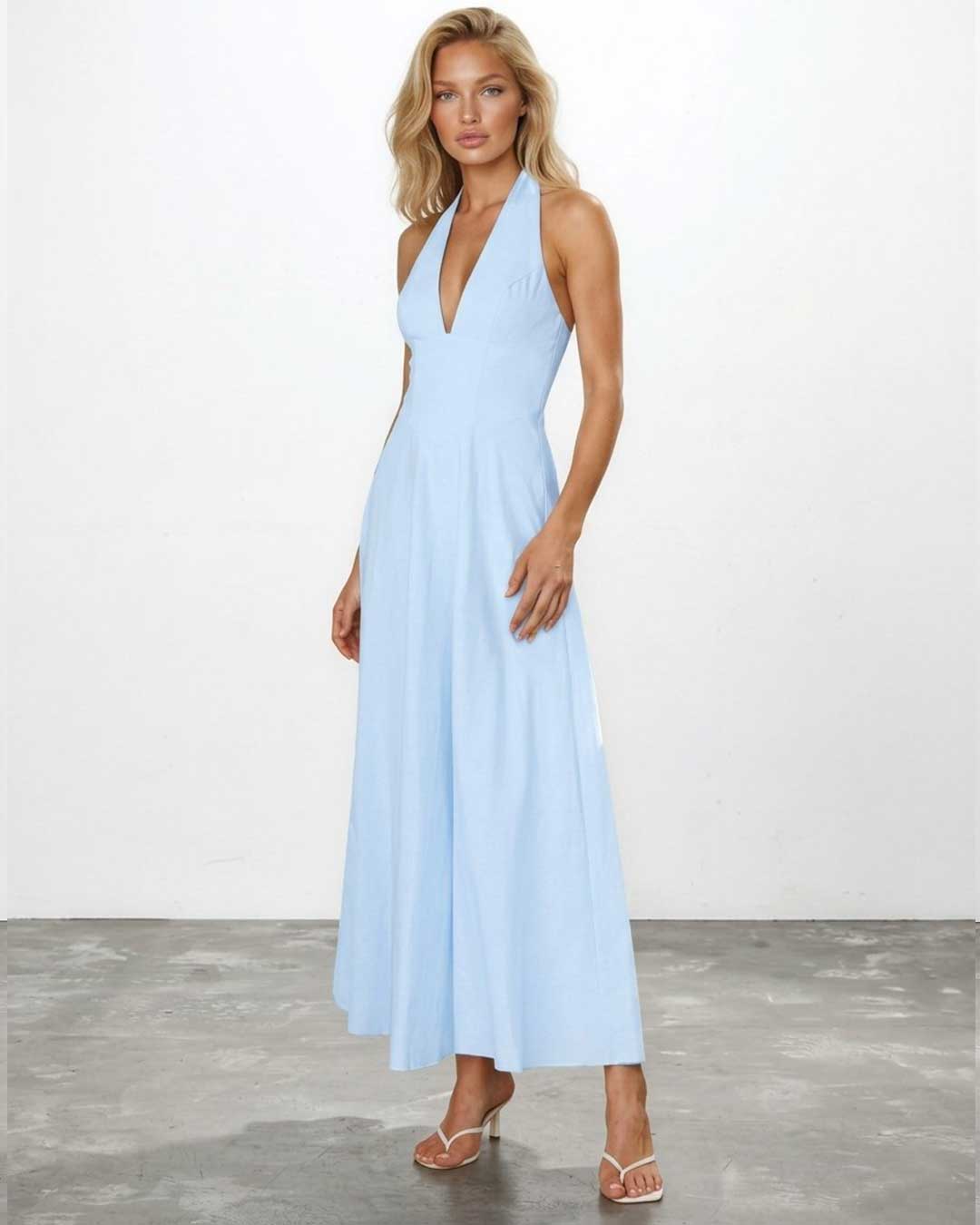 A woman modeling a light blue halter-neck maxi dress with a deep V-neckline, photographed against a white studio wall and gray floor.