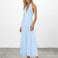 A woman modeling a light blue halter-neck maxi dress with a deep V-neckline, photographed against a white studio wall and gray floor.