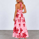 A woman modeling a pink halter-neck maxi dress with a bold red floral print, shown from the back against a light gray studio background.