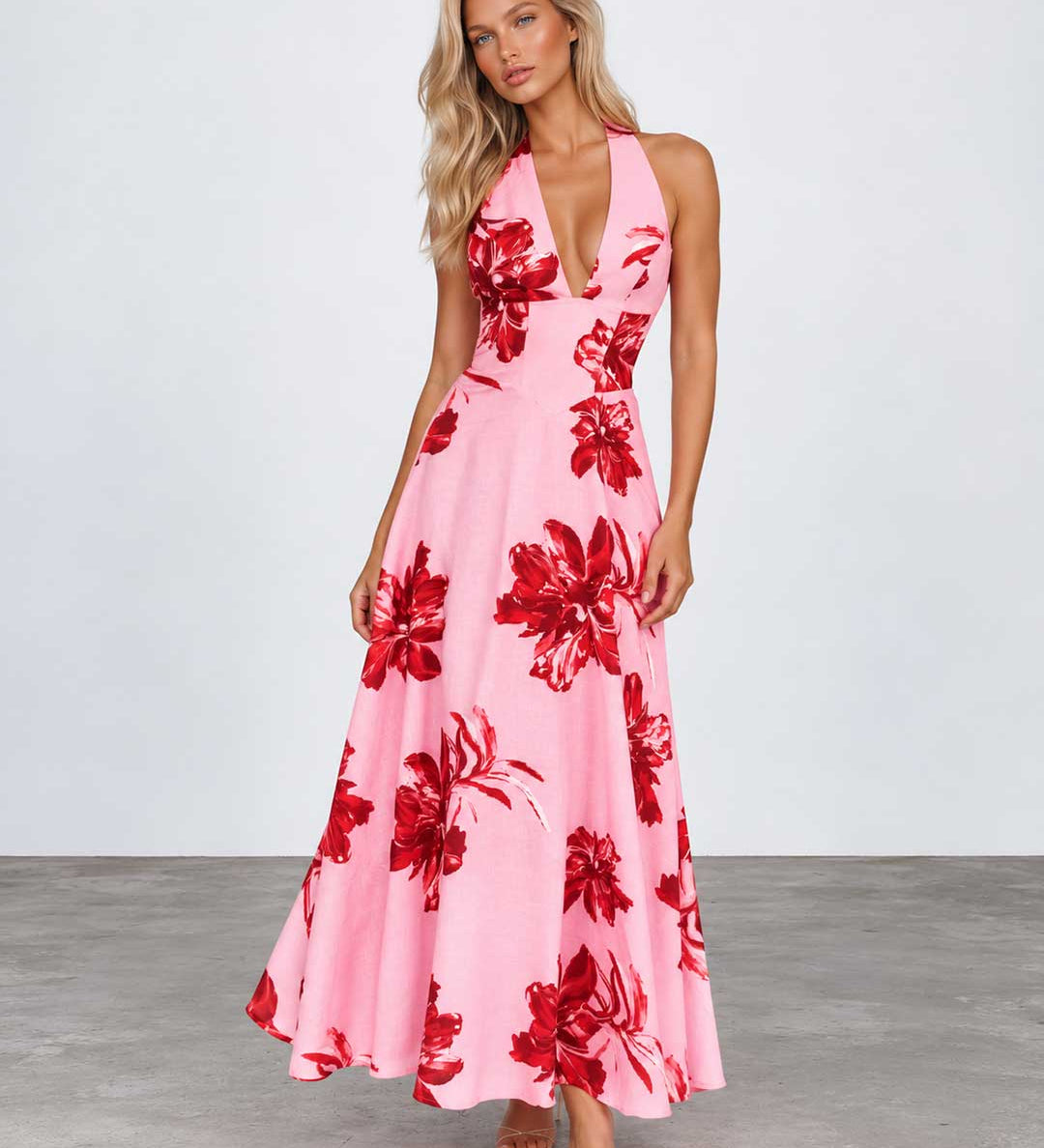 A woman modeling a pink halter-neck maxi dress with a bold red floral print, standing against a light gray studio background.