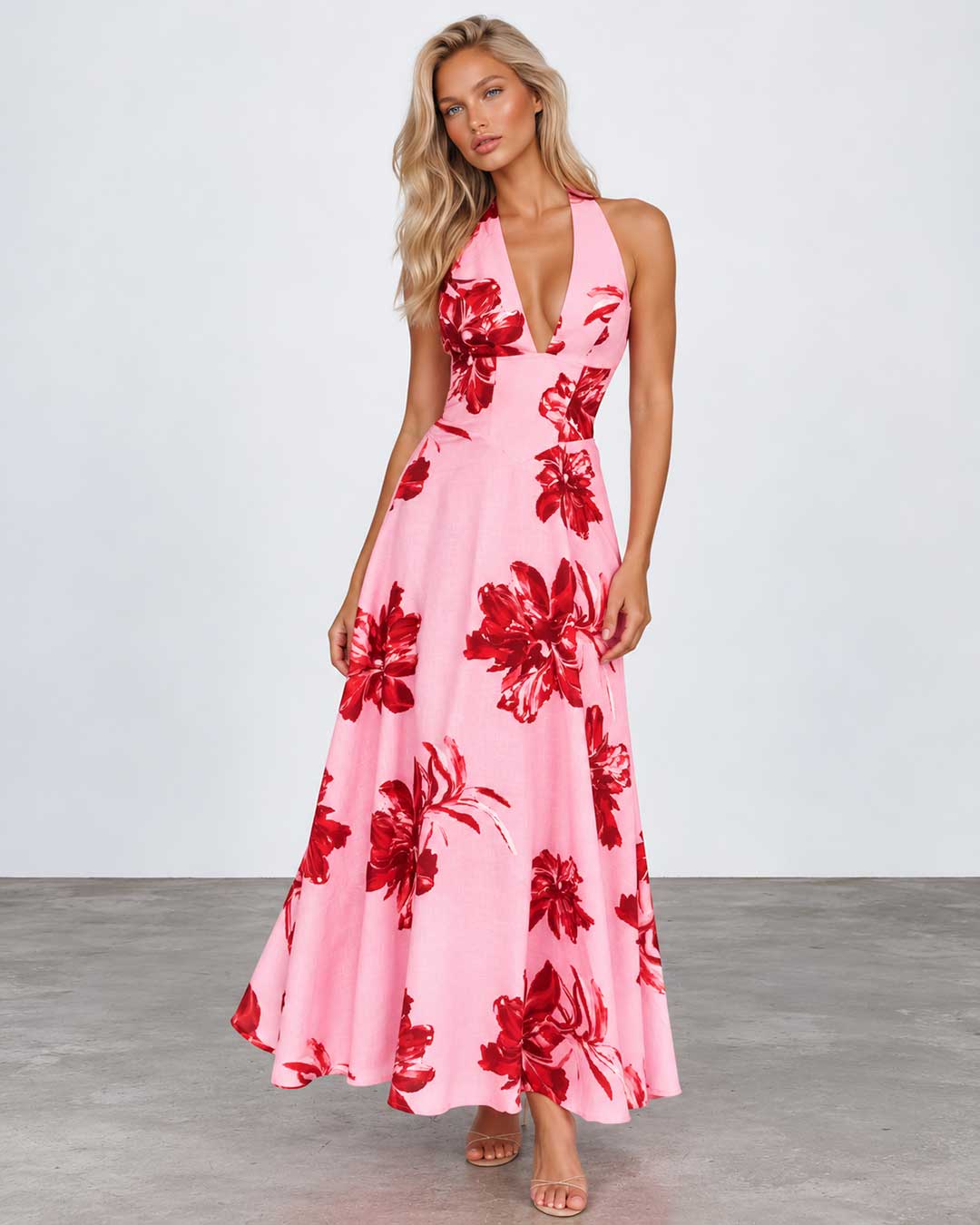 A woman modeling a pink halter-neck maxi dress with a bold red floral print, standing against a light gray studio background.