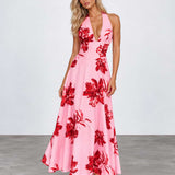 A woman modeling a pink halter-neck maxi dress with a bold red floral print, standing against a light gray studio background.