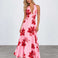 A woman modeling a pink halter-neck maxi dress with a bold red floral print, standing against a light gray studio background.