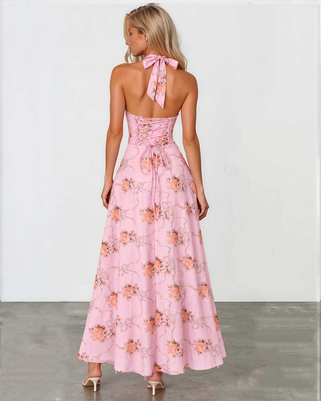 A woman wearing a pink halter-neck maxi dress with a floral print, shown from the back against a white studio wall and gray floor.