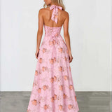 A woman wearing a pink halter-neck maxi dress with a floral print, shown from the back against a white studio wall and gray floor.