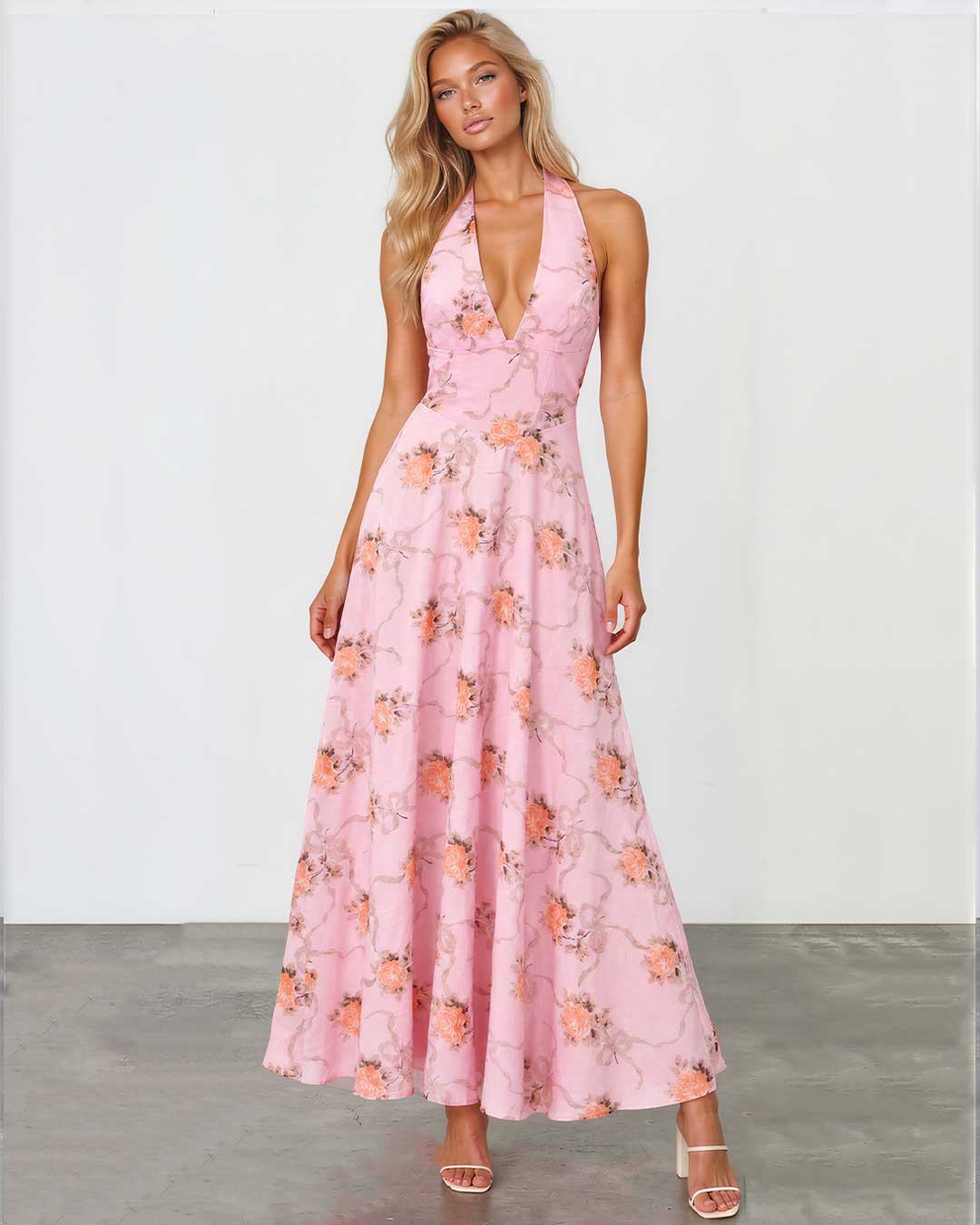 A woman modeling a pink halter-neck maxi dress with a floral print, standing against a white studio wall and gray floor.