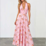 A woman modeling a pink halter-neck maxi dress with a floral print, standing against a white studio wall and gray floor.