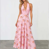 A woman modeling a pink halter-neck maxi dress with a floral print, standing against a white studio wall and gray floor.