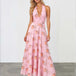 A woman modeling a pink halter-neck maxi dress with a floral print, standing against a white studio wall and gray floor.