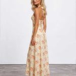 A woman modeling a sleeveless maxi dress with a floral print in beige, orange, and pink, shown from a side-back angle against a white studio wall.