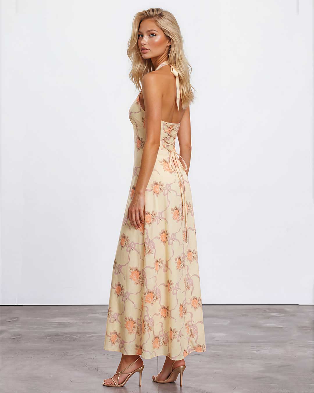 A woman modeling a sleeveless maxi dress with a floral print in beige, orange, and pink, shown from a side-back angle against a white studio wall.