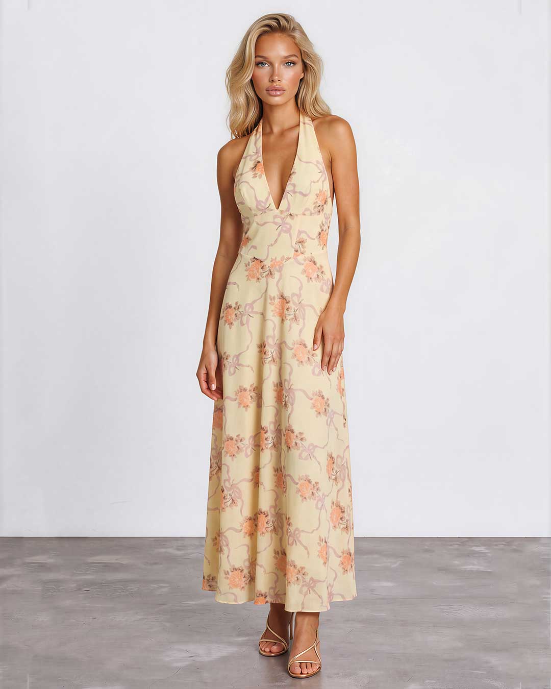 A woman modeling a sleeveless halter-neck maxi dress with a deep V-neckline, featuring a light beige base with a soft peach and pink floral pattern, set against a plain light gray studio background.