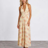 A woman modeling a sleeveless halter-neck maxi dress with a deep V-neckline, featuring a light beige base with a soft peach and pink floral pattern, set against a plain light gray studio background.