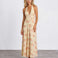 A woman modeling a sleeveless halter-neck maxi dress with a deep V-neckline, featuring a light beige base with a soft peach and pink floral pattern, set against a plain light gray studio background.