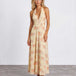 A woman modeling a sleeveless halter-neck maxi dress with a deep V-neckline, featuring a light beige base with a soft peach and pink floral pattern, set against a plain light gray studio background.