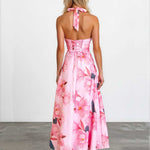 A woman viewed from behind wearing a pink floral halter-neck maxi dress with lace-up back detailing, standing against a plain white wall on a gray floor.