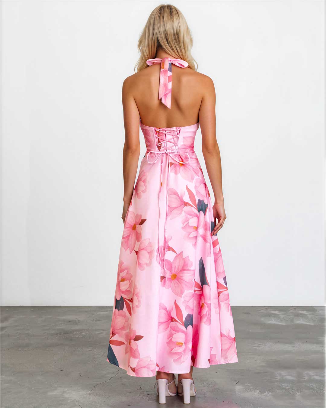 A woman viewed from behind wearing a pink floral halter-neck maxi dress with lace-up back detailing, standing against a plain white wall on a gray floor.