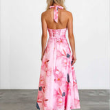 A woman viewed from behind wearing a pink floral halter-neck maxi dress with lace-up back detailing, standing against a plain white wall on a gray floor.