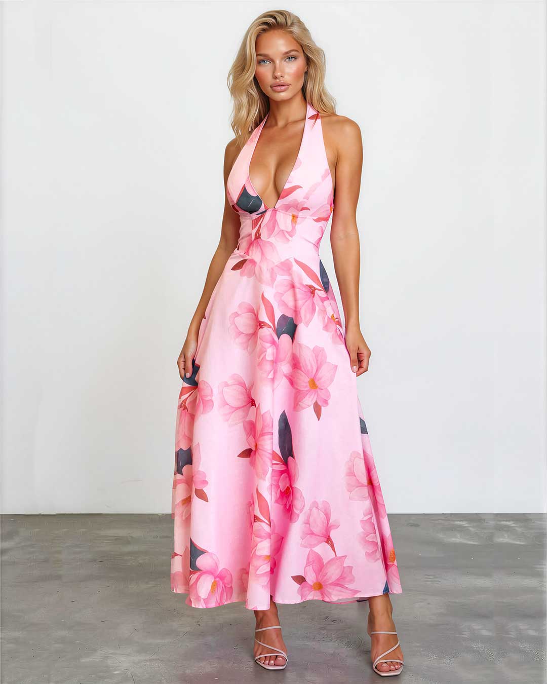 A woman modeling a pink halter-neck maxi dress with a large floral print in pink, red, and dark gray, standing against a white studio wall.