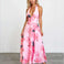 A woman modeling a pink halter-neck maxi dress with a large floral print in pink, red, and dark gray, standing against a white studio wall.