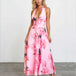 A woman modeling a pink halter-neck maxi dress with a large floral print in pink, red, and dark gray, standing against a white studio wall.