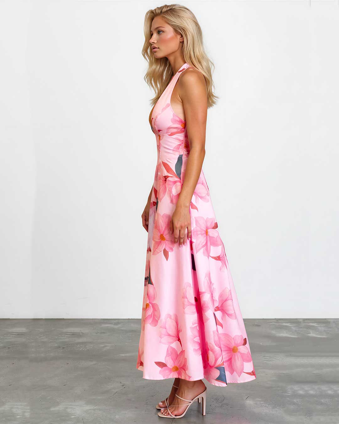 A woman modeling a pink halter-neck maxi dress with a large floral print, standing in profile against a white wall and gray floor.