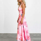 A woman modeling a pink halter-neck maxi dress with a large floral print, standing in profile against a white wall and gray floor.