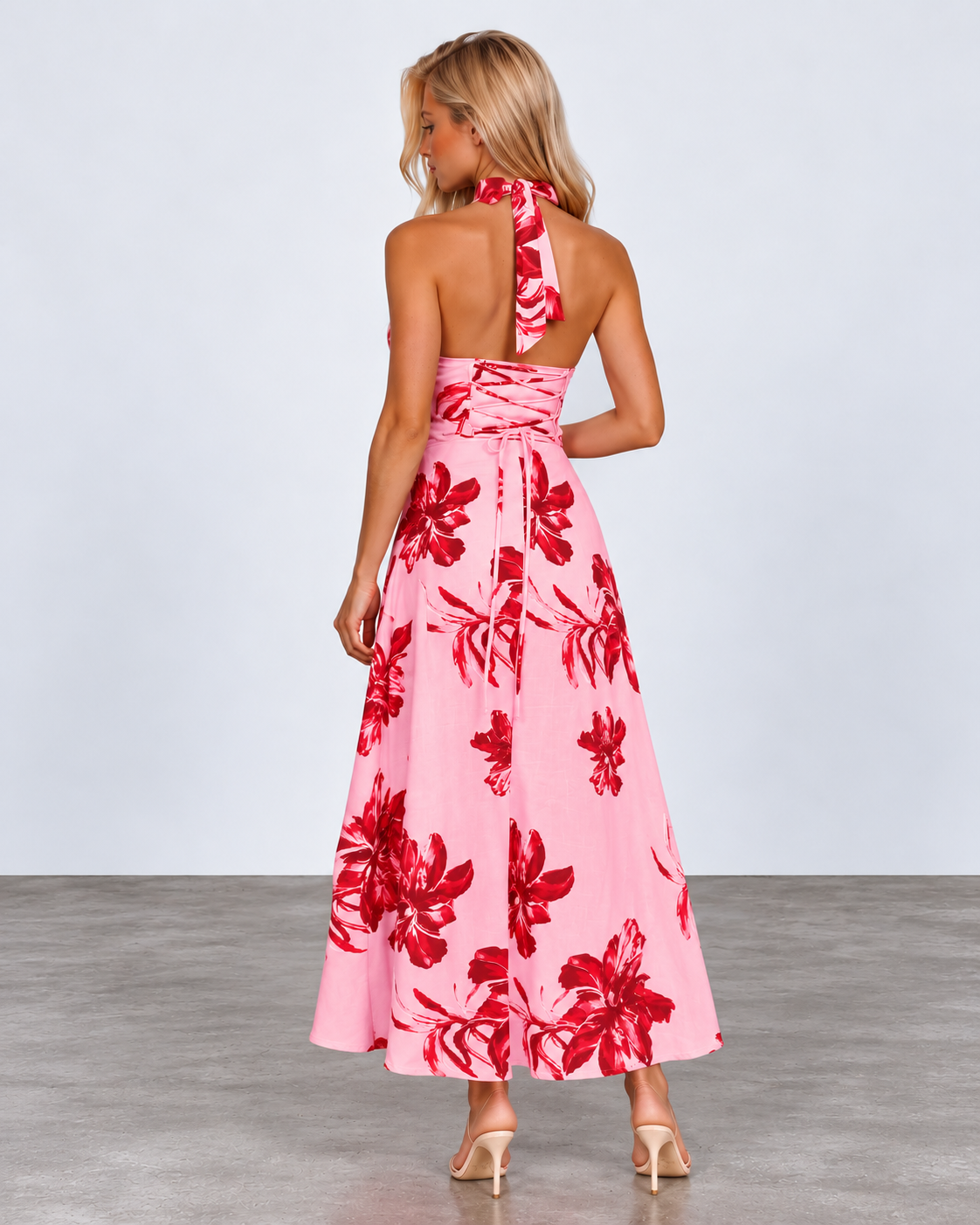 A woman modeling a pink halter-neck maxi dress with a bold red floral pattern, shown from the back against a light gray studio background.