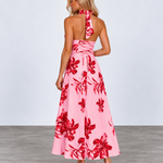 A woman modeling a pink halter-neck maxi dress with a bold red floral pattern, shown from the back against a light gray studio background.