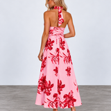 A woman modeling a pink halter-neck maxi dress with a bold red floral pattern, shown from the back against a light gray studio background.