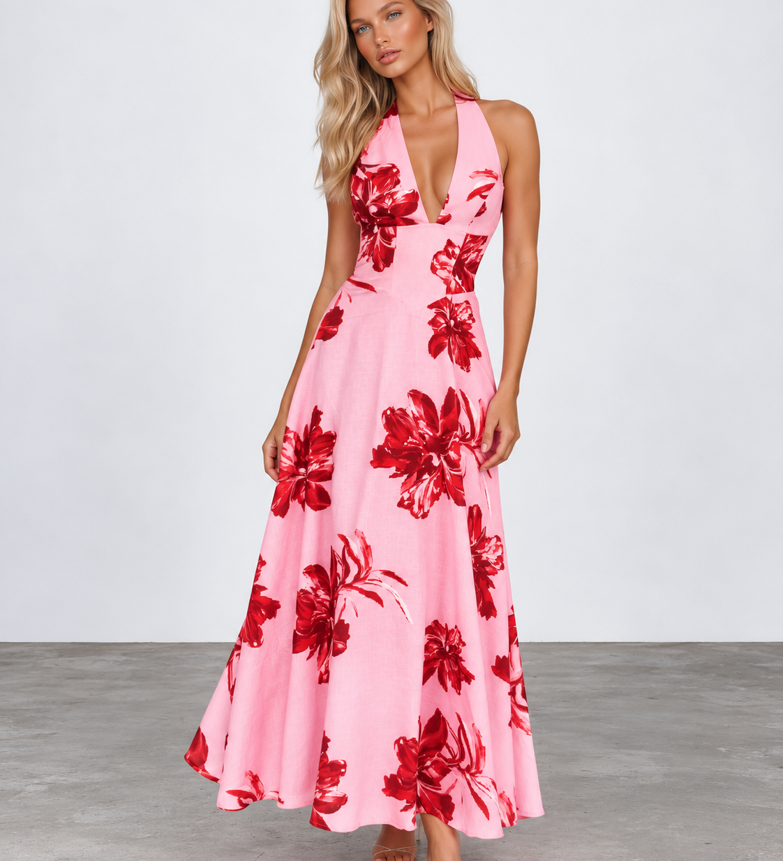 A woman modeling a pink halter-neck maxi dress with a bold red floral print, standing against a light gray studio background.