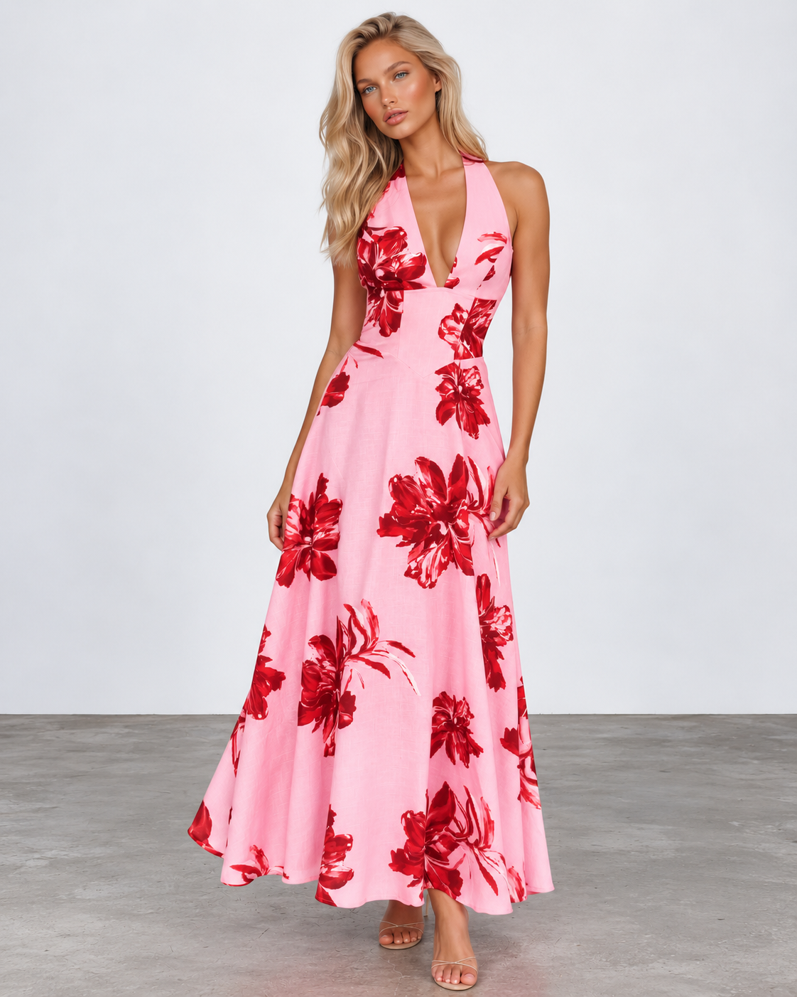 A woman modeling a pink halter-neck maxi dress with a bold red floral print, standing against a light gray studio background.