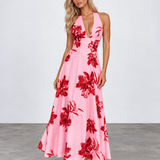A woman modeling a pink halter-neck maxi dress with a bold red floral print, standing against a light gray studio background.