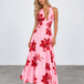 A woman modeling a pink halter-neck maxi dress with a bold red floral print, standing against a light gray studio background.