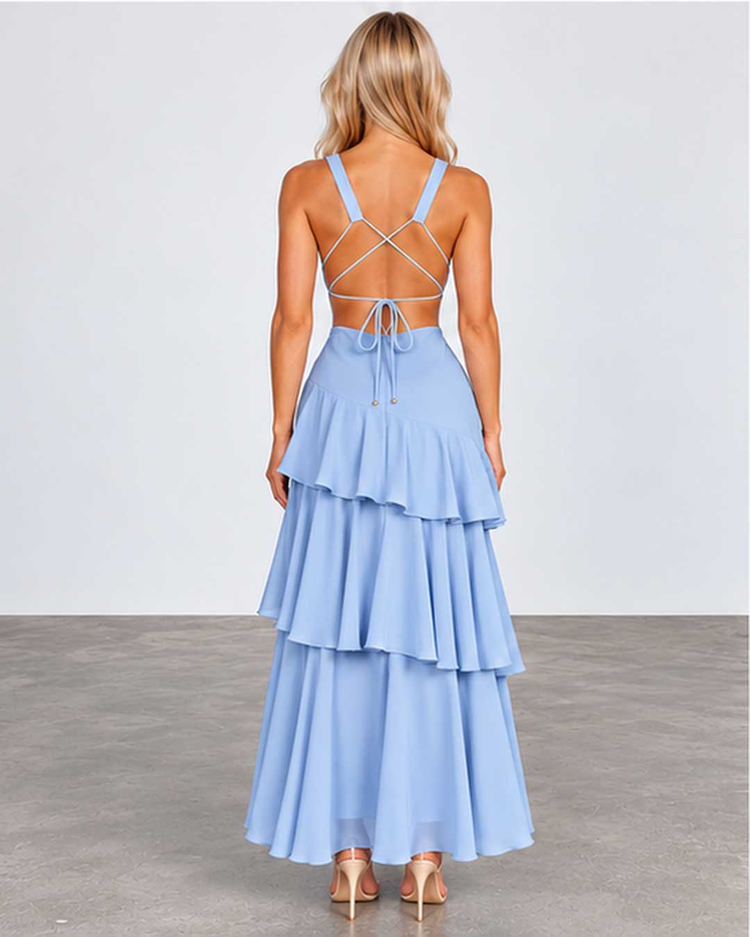 A woman viewed from the back wearing a light blue sleeveless maxi dress with a strappy crisscross open back and a tiered ruffled skirt, paired with nude heels, against a plain studio backdrop.