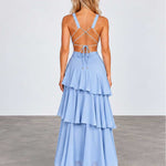 A woman viewed from the back wearing a light blue sleeveless maxi dress with a strappy crisscross open back and a tiered ruffled skirt, paired with nude heels, against a plain studio backdrop.