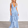A woman modeling a light blue sleeveless maxi dress with a deep V-neckline and a tiered ruffle skirt, standing against a neutral studio backdrop.