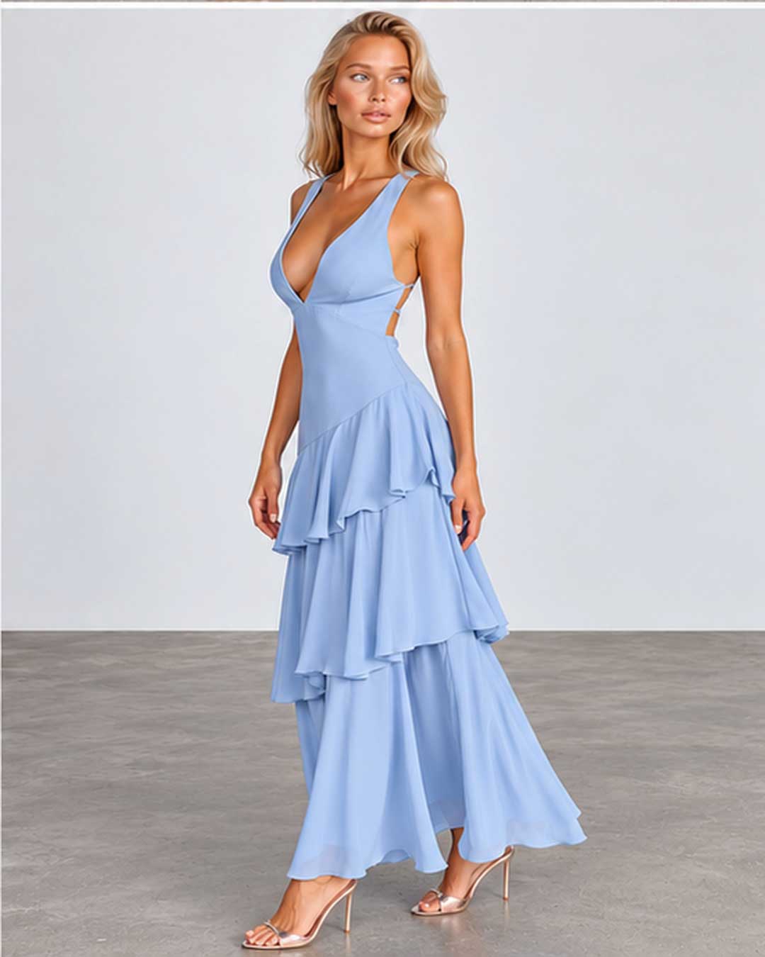 A woman modeling a light blue sleeveless maxi dress with a deep V-neckline, side cutouts, and a tiered ruffled skirt, paired with clear high-heeled sandals against a neutral studio backdrop.
