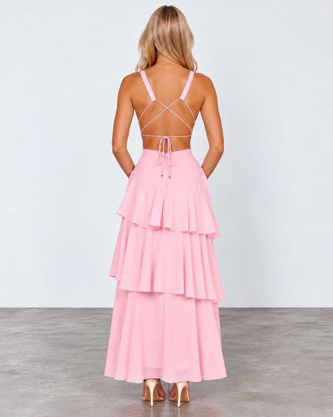 A woman stands with her back to the camera wearing a light pink sleeveless maxi dress with a strappy open back and a tiered ruffle skirt, paired with nude heels, in a studio setting.