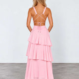 A woman stands with her back to the camera wearing a light pink sleeveless maxi dress with a strappy open back and a tiered ruffle skirt, paired with nude heels, in a studio setting.