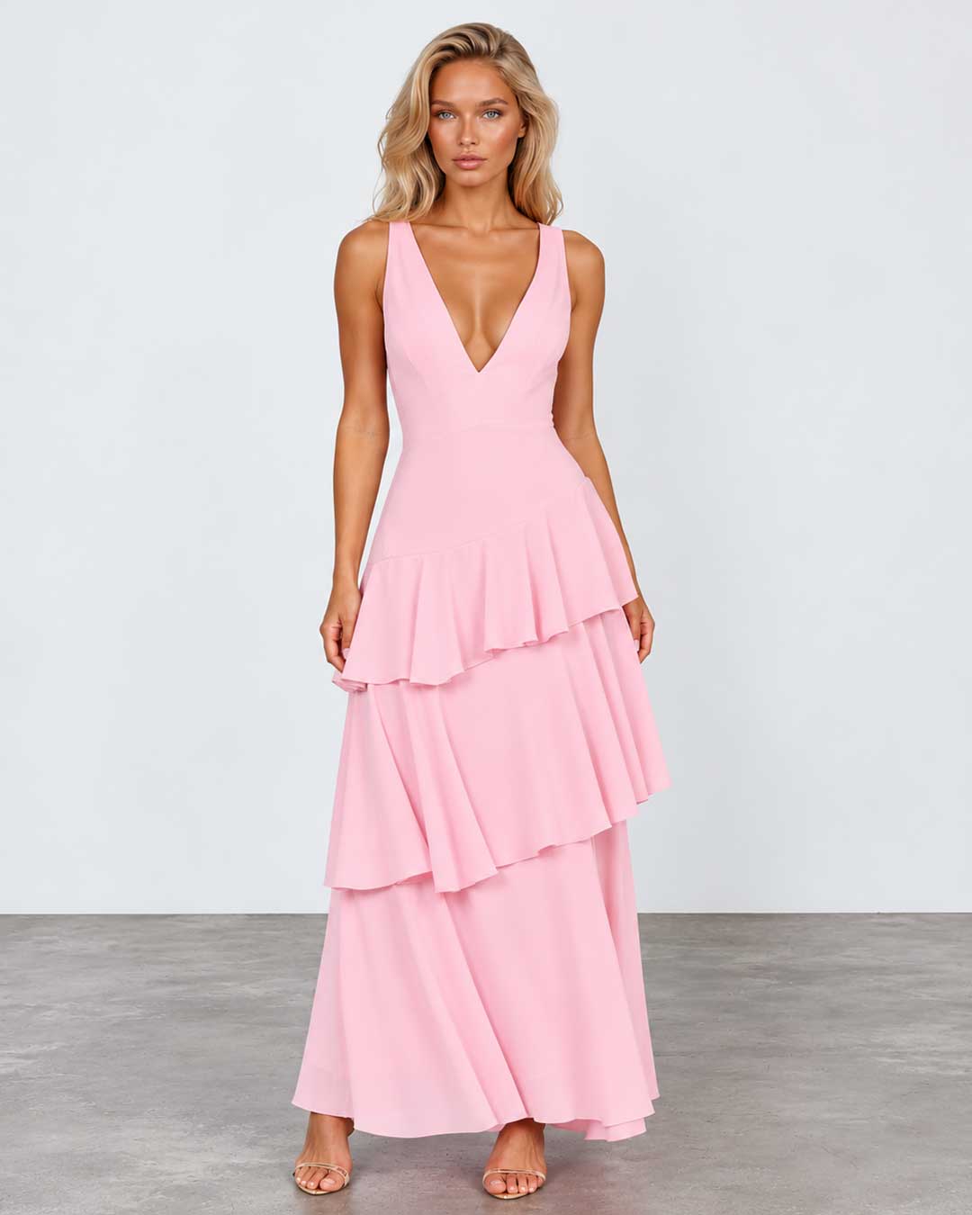 A woman modeling a light pink sleeveless maxi dress with a deep V-neckline and a tiered ruffle skirt, standing against a light gray studio background.