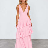 A woman modeling a light pink sleeveless maxi dress with a deep V-neckline and a tiered ruffle skirt, standing against a light gray studio background.