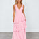 A woman modeling a light pink sleeveless maxi dress with a deep V-neckline and a tiered ruffle skirt, standing against a light gray studio background.