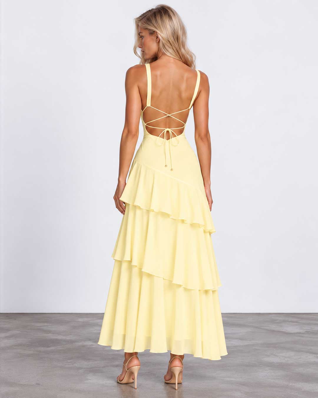 A woman modeling a pale yellow tiered maxi dress from the back, featuring thin straps, a strappy open back, and a flowing layered skirt, set against a plain studio background.
