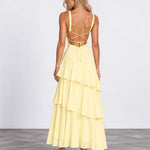 A woman modeling a pale yellow tiered maxi dress from the back, featuring thin straps, a strappy open back, and a flowing layered skirt, set against a plain studio background.