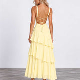 A woman modeling a pale yellow tiered maxi dress from the back, featuring thin straps, a strappy open back, and a flowing layered skirt, set against a plain studio background.