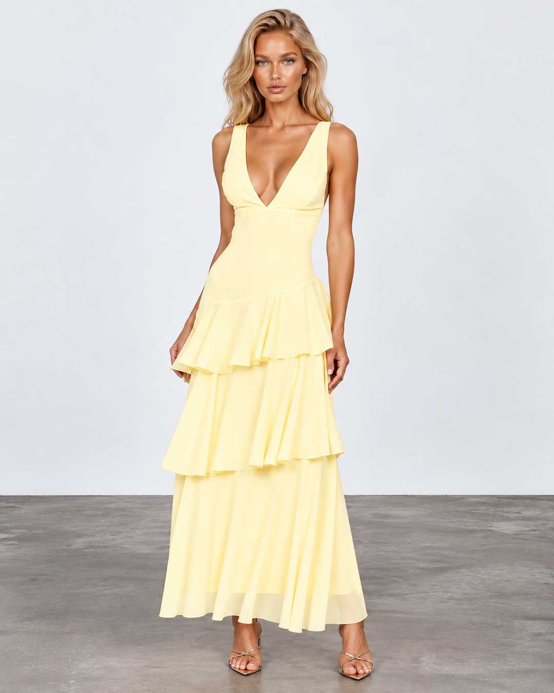 A woman modeling a pale yellow sleeveless maxi dress with a deep V-neckline, fitted bodice, and tiered ruffle skirt, standing against a light gray studio background.